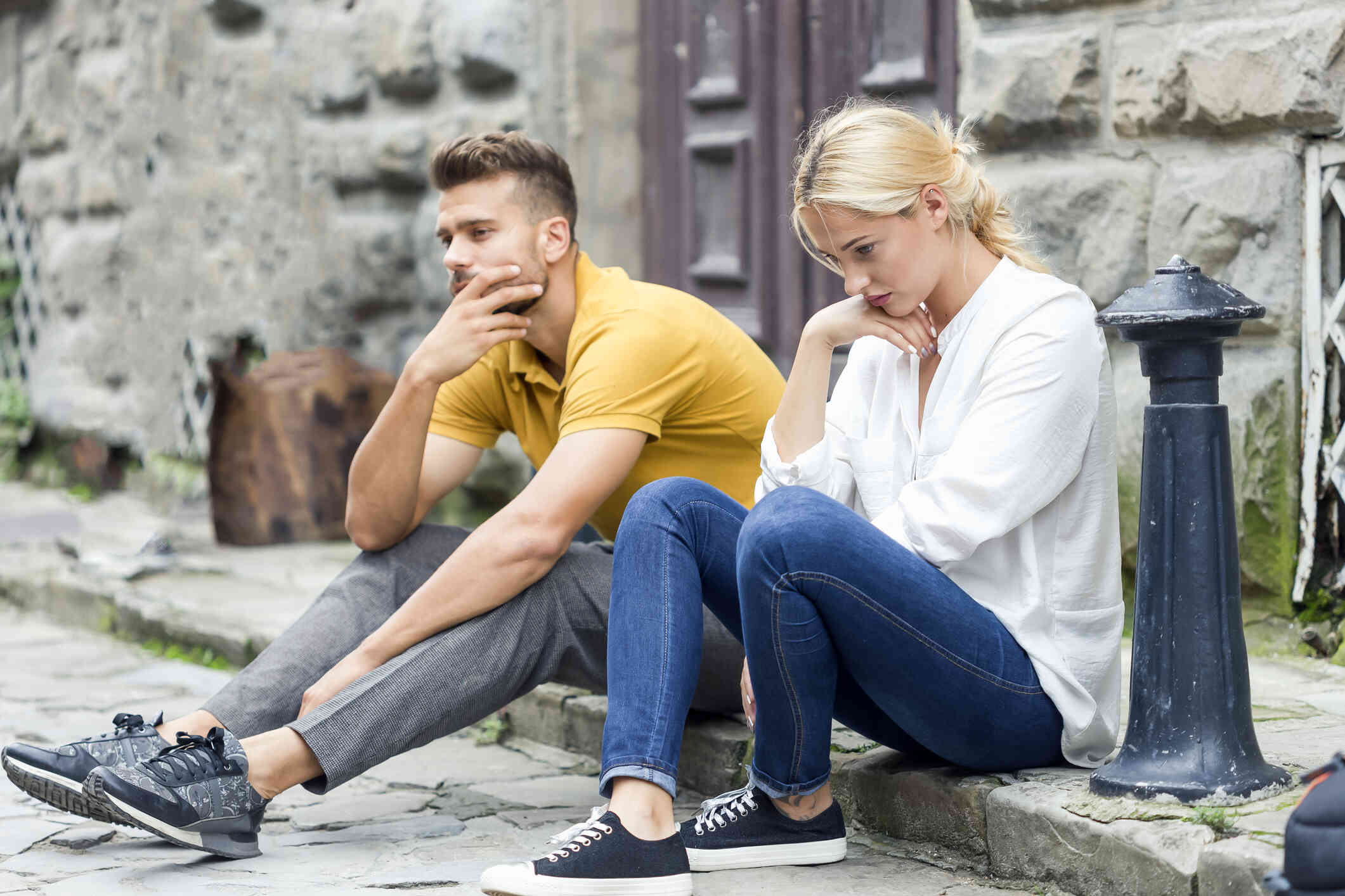 A male and female couple sit next to each other on a step outside a building while gazing down sadly.